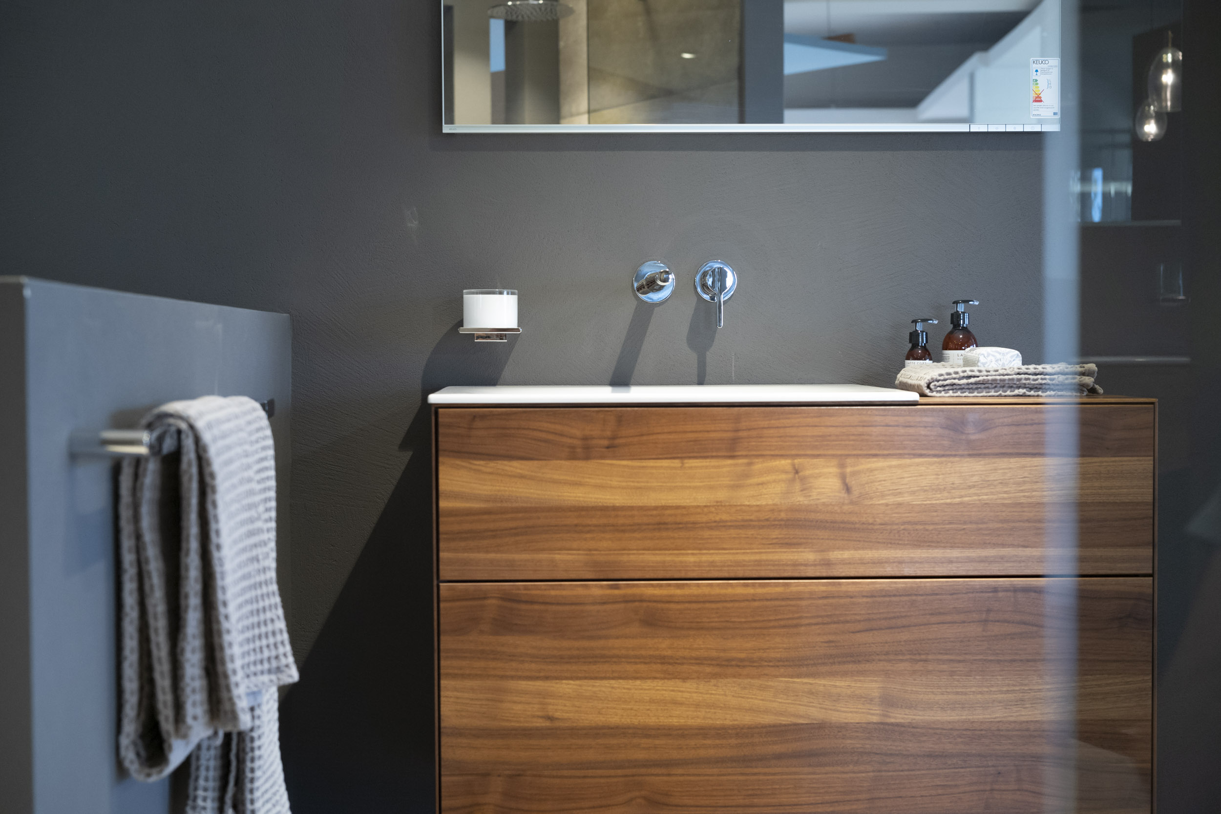 Bad-Ausstellung bei Sanitär Bez: moderner Waschtisch mit warm gemaserter Holzfront und zwei breiten Schubladen, darauf eine weiße, flache Waschtischplatte. An der dunkelgrauen Wand darüber sind ein wandmontierter Chrom-Auslauf und ein runder Bediengriff zu sehen, oben schneidet ein Spiegel ins Bild. Rechts liegen gefaltete Handtücher mit zwei braunen Seifenspendern, links im Vordergrund hängt ein graues Handtuch an einer Halterung; eine Glasreflexion verläuft als helle vertikale Linie durch das Motiv.