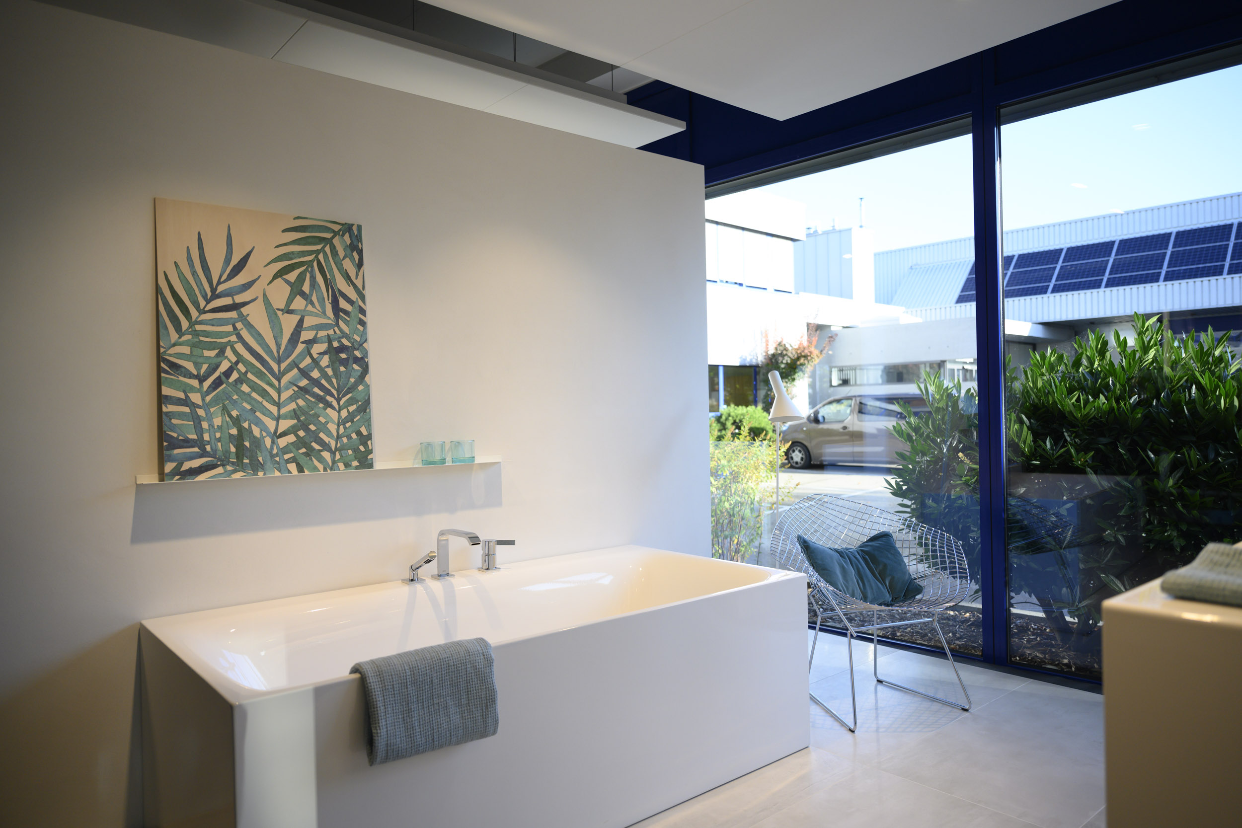 Heller Bad-Ausstellungsraum bei Sanitär Bez mit einer freistehenden, rechteckigen Badewanne in glänzendem Weiß und verchromter Wannenarmatur; über der Wanne hängt ein großes Bild mit Blattmuster und darunter ein schmales Wandboard mit zwei hellblauen Gläsern. Rechts sorgt eine bodentiefe Fensterfront mit dunkelblauem Rahmen für viel Tageslicht und Blick ins Freie auf Grünpflanzen, einen Parkplatzbereich und Gebäude mit Solarpanels. Vor dem Fenster steht ein filigraner Drahtstuhl mit blauem Kissen, ein graues Handtuch hängt über dem Wannenrand, und der Raum wirkt minimalistisch, ruhig und modern.